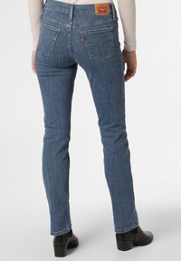 Blue denim jeans with a slim fit, featuring a mid-rise waist, rear pockets, and a leather patch at the back waist. Smooth texture.