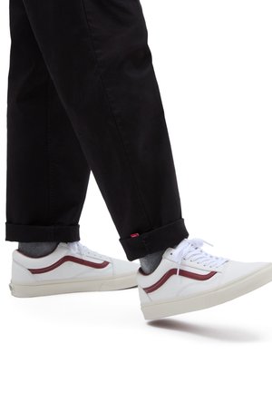 Personne portant un pantalon noir retroussé, des chaussettes grises et des baskets blanches avec une bande rouge marchant sur un fond blanc.