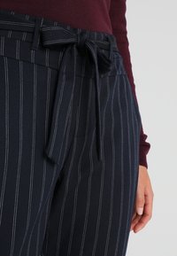 Granatowe spodnie w białe pionowe paski, z paskiem zawiązanym w talii i gładką fakturą. Materiał wydaje się solidny i dobrze skrojony.