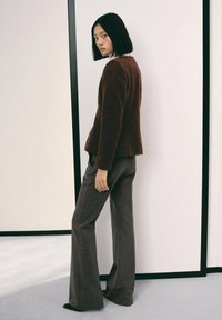 Veste marron texturée associée à un pantalon large gris. L'ensemble présente un design simple et épuré avec des accents minimaux.