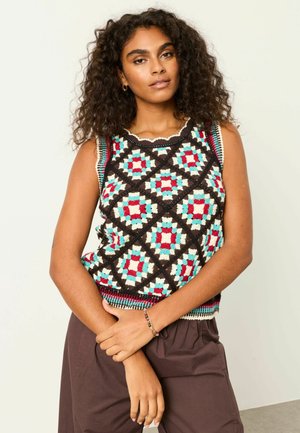 Frau mit lockigem Haar, die ein farbenfrohes, gehäkeltes ärmelloses Oberteil mit geometrischen Mustern und braune Hosen trägt, vor einem einfachen Hintergrund stehend.