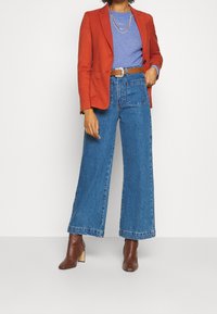 Rode blazer, blauwe gebreide top, wijdvallende blauwe jeans en bruine haklaarzen. Opvallende textuur op de stof en gelaagd kettingdetail.