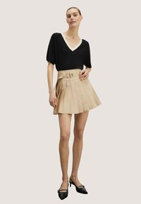 Top nero a V con maniche corte, abbinato a una gonna plissettata beige con cintura in vita. Tacchi neri completano l'outfit.