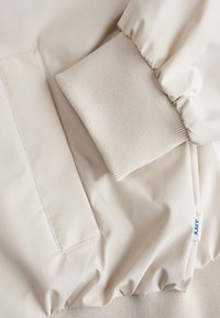Detalje af beige jakkeærme og lomme med ribbet manchet og elastisk linning med lille stofstørrelsesmærkat.