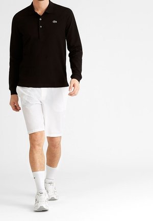 Mann trägt schwarzen langärmligen Poloshirt, weiße Shorts, weiße Socken und weiße Turnschuhe und geht vor einem schlichten weißen Hintergrund.