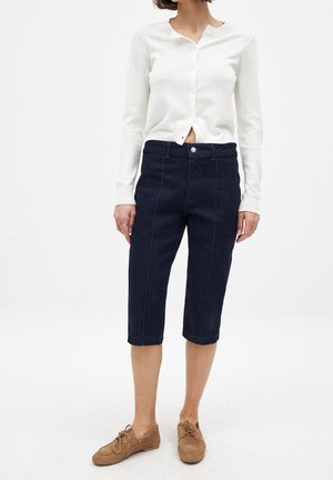 Biely dlhý cardigan s dlhými rukávmi a tmavomodré skrátené denimsové nohavice s prednými švami. Hnedé šnurovacie topánky. Minimalistický dizajn, priliehavý silueta.