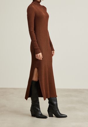 Femme portant une robe en maille côtelée marron à manches longues avec une fente latérale et des bottes noires à talons mi-mollet sur un fond clair.
