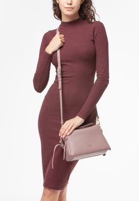 Borsa a tracolla in pelle rosa chiaro con finitura testurizzata, dettagli in metallo dorato e tracolla regolabile, tenuta accanto a una figura in un vestito bordeaux.
