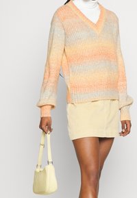 Pull en maille à col en V dégradé dans des teintes d'orange et de jaune. Présentant une texture côtelée et des manches amples. Associé à une jupe en velours côtelé de couleur claire et un sac à main jaune.
