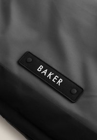 Čierna taška z textilu s hladkým povrchom, obsahujúca obdĺžnikovú tkanú nálepku s nápisom "BAKER" v bielej farbe. Detaily gombíka na nálepke.