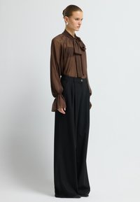 Braune, durchsichtige Bluse mit Halstuch und weit ausgestellten Ärmeln, kombiniert mit hochgeschnittenen, schwarzen, weit geschnittenen Hosen mit Knopfdetails vorne.