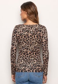 Frau mit langen braunen Haaren, die ein langärmliges Oberteil mit Leopardenmuster und blaue Jeans trägt, steht vor einer hellen Wand, Rücken sichtbar.
