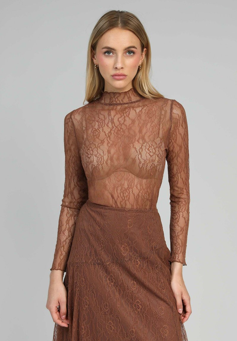 Top en dentelle marron à manches longues et col haut, avec des motifs floraux complexes et une coupe ajustée. Le matériau transparent souligne les caractéristiques.