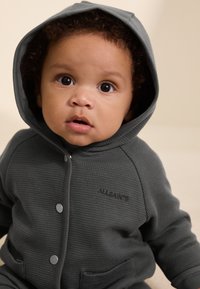 Gray hooded onesie made of textured fabric, featuring snap buttons and front pockets. Logo displayed on the chest.