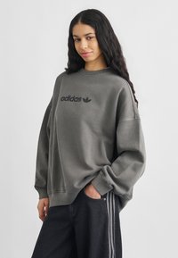 Νέα γυναίκα με μακριά σκούρα μαλλιά που φοράει γκρι φούτερ Adidas και σκούρο τζιν με άσπρες ρίγες στο πλάι, στέκεται με το ένα χέρι στην τσέπη.