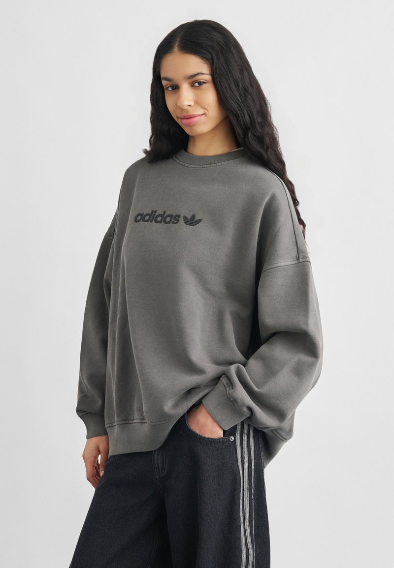 Νέα γυναίκα με μακριά σκούρα μαλλιά που φοράει γκρι φούτερ Adidas και σκούρο τζιν με άσπρες ρίγες στο πλάι, στέκεται με το ένα χέρι στην τσέπη.