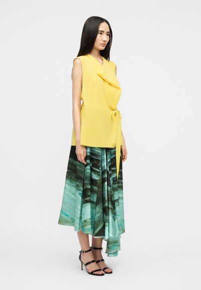 Femme portant un haut cache-cœur jaune sans manches avec ceinture nouée à la taille et une jupe midi fluide à motifs verts et bleus, avec des talons noirs à brides, debout de profil.