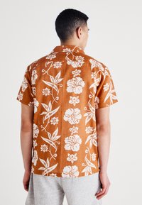 Camisa de manga corta marrón con estampado floral, que presenta un patrón floral blanco, diseño con collar y un corte relajado, hecha de una tela texturizada.