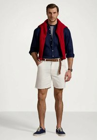 Moški v mornarsko modri srajci čez črtasto majico, rdeč pulover čez ramena, bele kratke hlače, pleteni pas in mornarsko modre superge, stoji z rokami v žepih.