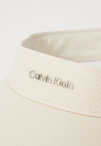 Calvin Klein solhatt i kremet beige stoff med diagonalt ribbet tekstur. Den har en sømmet rund kant og en sølvlogo på forsiden.