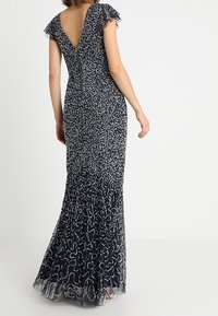 Námořnická modrá večerní šaty s intricátními stříbrnými korálky, jemnými rukávy, V-zadním výstřihem a splývavým texturovaným lemem s průsvitným překryvem.