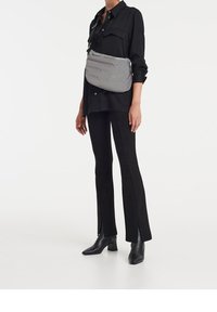 Borsa a tracolla grigia imbottita con una texture liscia, dotata di chiusura zip superiore e tracolla regolabile, indossata sopra una camicia nera oversize e pantaloni.