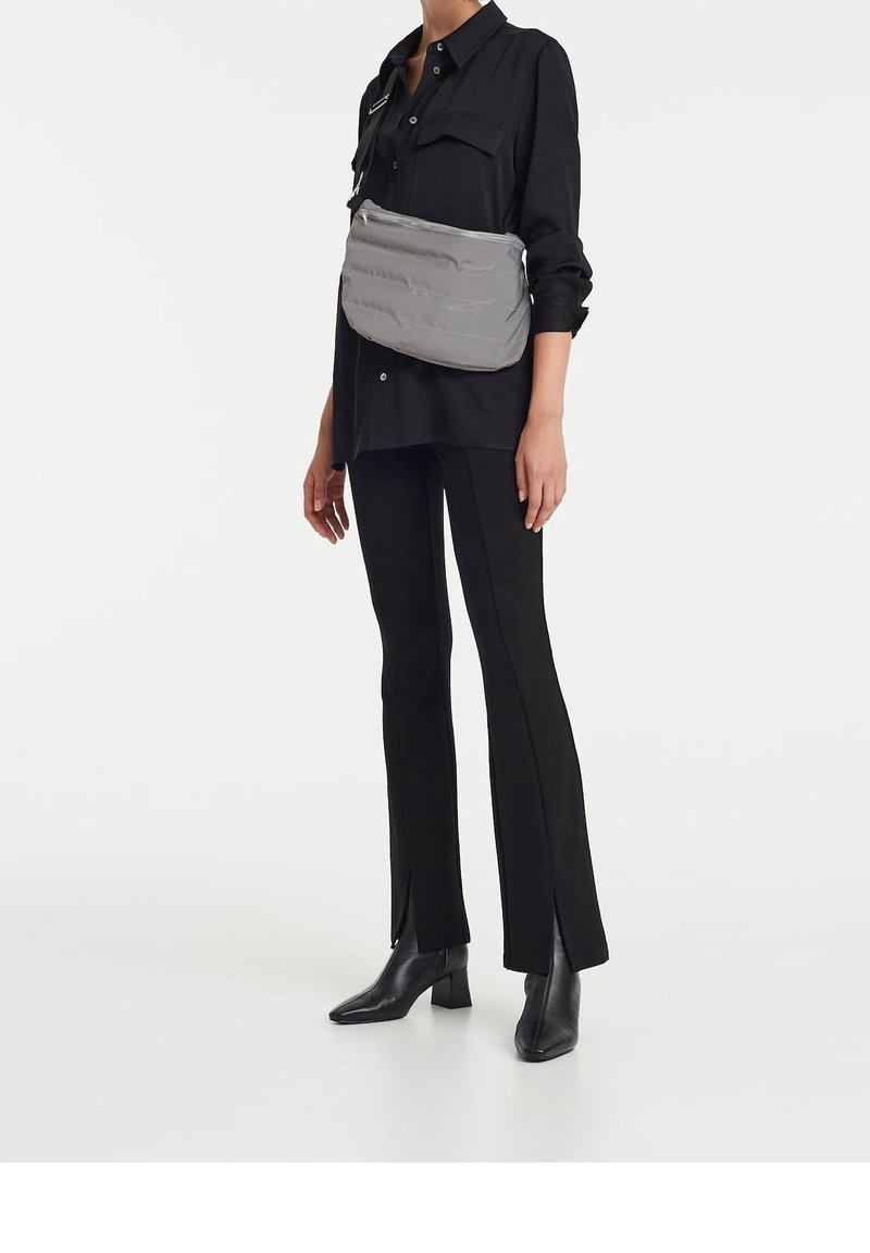 Borsa a tracolla grigia imbottita con una texture liscia, dotata di chiusura zip superiore e tracolla regolabile, indossata sopra una camicia nera oversize e pantaloni.