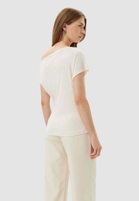 Top bianco a maniche corte con vestibilità ampia e design scollato sulle spalle. Texture liscia, dettagli minimi, abbinato a pantaloni chiari.