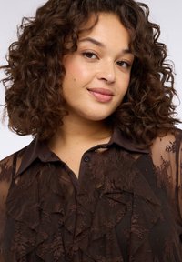 Camicia marrone trasparente con dettagli in pizzo stratificati, colletto e chiusura con bottoni. Tessuto strutturato con motivi arricciati e design aderente.