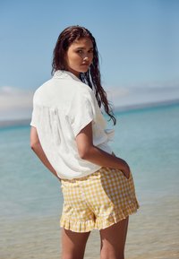 Hvid hørskjorte med knapper og opkrogede ærmer, kombineret med gule ternede shorts med flæser. Baggrunden viser en strand og blå himmel.