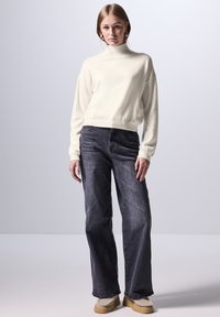 Maglione a collo alto di colore crema con polsini a coste, abbinato a jeans a gamba larga in denim scuro. Il modello indossa scarpe slip-on beige con rivestimento in pelo.