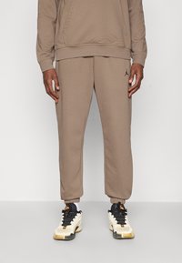 Brune bomullsblanding joggebukser med elastiske mansjetter, med en svart logo på venstre lår og en avslappet passform, kombinert med svart-hvite sneakers.