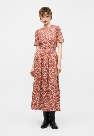 Γυναίκα που στέκεται φορώντας κόκκινο φόρεμα με κοντά μανίκια και σχέδιο με ανθάκια, με ζώνη στη μέση και μαύρες μπότες μέχρι τη μέση της γάμπας, σε απλό φόντο.