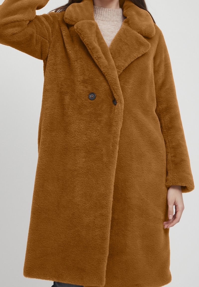 Femme portant un manteau marron à double boutonnage en fausse fourrure sur un pull beige clair, sur un fond uni.
