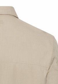 Beiges Hemd mit verbreitetem Kragen, aus strukturiertem Stoff mit feinem Glanz. Nahtdetails sind nahe am Kragen und an der Schulter sichtbar.