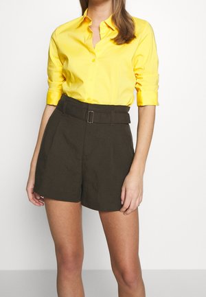Kvinde iført en klar gul skjorte med knapper og op rullede ærmer samt højtaljede mørkebrune shorts med bælte.