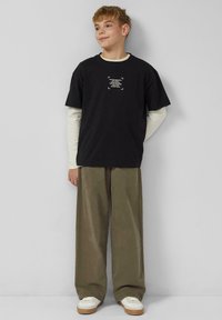 Zwart katoenen T-shirt met witte tekstprint, gedragen over een crèmekleurig shirt met lange mouwen. Losse olijfgroene broek en witte sneakers.