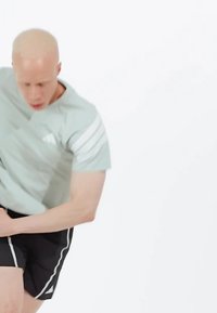 Camiseta atlética verde claro con tres franjas blancas en el hombro, combinada con pantalones cortos negros que tienen franjas blancas en los laterales. Pose dinámica.