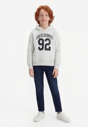 Κοκκινομάλλης αγόρι που φοράει ανοιχτό γκρι φούτερ Abercrombie με το νούμερο "92", σκούρα μπλε τζιν, γκρι κάλτσες και λευκά αθλητικά παπούτσια, στέκεται μπροστά σε λευκό φόντο.