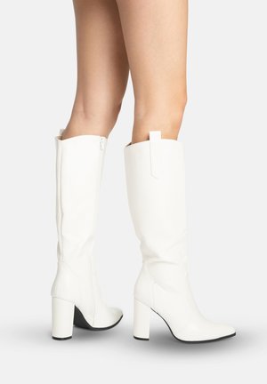 Stivali bianchi a metà polpaccio con tacco e cerniera laterale indossati da una persona in piedi su una superficie bianca su uno sfondo semplice.