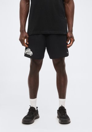 Άνδρας με μαύρο αθλητικό σορτς με λευκό σχέδιο στο αριστερό πόδι, μαύρα αθλητικά παπούτσια, λευκές κάλτσες και μαύρο μπλουζάκι σε φόντο απλό.