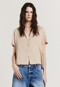 Blusa beige a maniche corte con chiusura frontale a bottoni, realizzata in tessuto liscio. Abbinata a jeans in denim blu chiaro a vita alta.