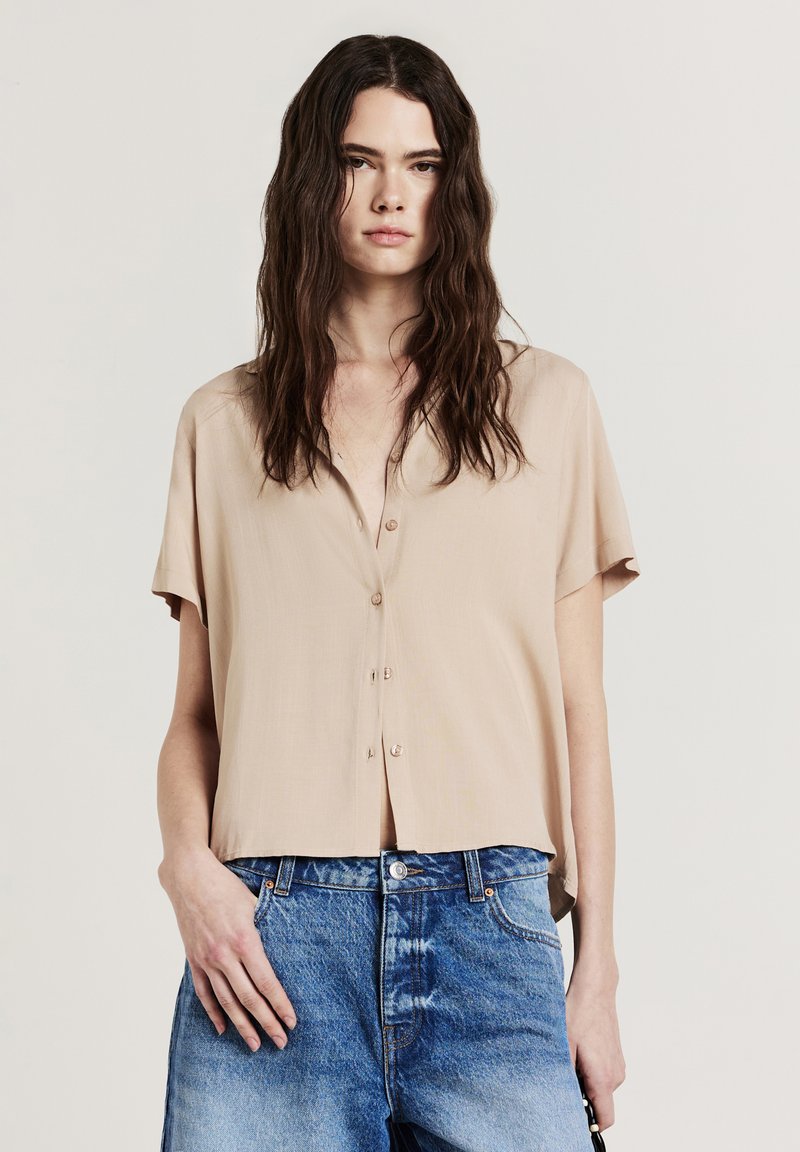 Blusa beige a maniche corte con chiusura frontale a bottoni, realizzata in tessuto liscio. Abbinata a jeans in denim blu chiaro a vita alta.
