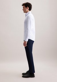 Witte lange mouwen shirt met een kraag, gecombineerd met marineblauwe broek en zwarte schoenen. Het shirt heeft een zijsplit en knopenmanchetten.