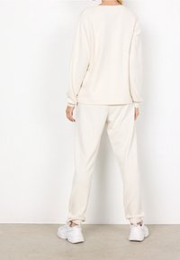 Mujer de pie de espaldas vistiendo una blusa de manga larga color blanco roto, pantalones jogger a juego con puños elásticos y zapatillas blancas sobre un suelo gris.