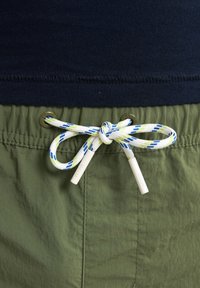 Pantaloni corti verdi in un tessuto testurizzato; presentano un cinturino blu navy e un cordoncino bianco con accenti blu e verdi, fissati da occhielli in metallo.