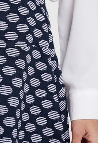 Vestido de punto con una base azul oscuro y círculos blancos de rayas horizontales. Incluye un primer plano de una camisa blanca de manga larga parcialmente visible.