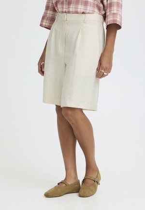 Žena nosi krémové šortky po kolená, ružovú kockovanú košeľu a olivovo zelené baleríny Mary Jane, stojaca so uvoľnenými rukami pozdĺž tela.