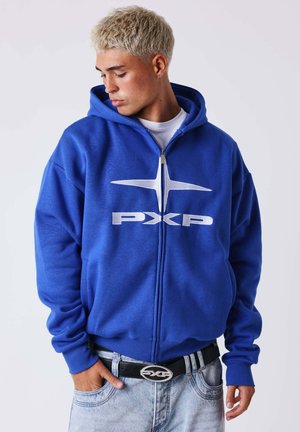 Jonge man met blond haar, gekleed in een blauwe hoodie met rits en een wit "PXP"-logo, lichtblauwe spijkerbroek en een zwarte riem met zilveren gesp.