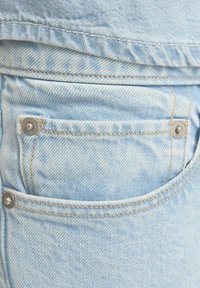 Unos jeans de mezclilla azul claro presentan un bolsillo trasero con remaches metálicos plateados y costuras en contraste en color marrón claro, destacando un acabado de tela texturizada.